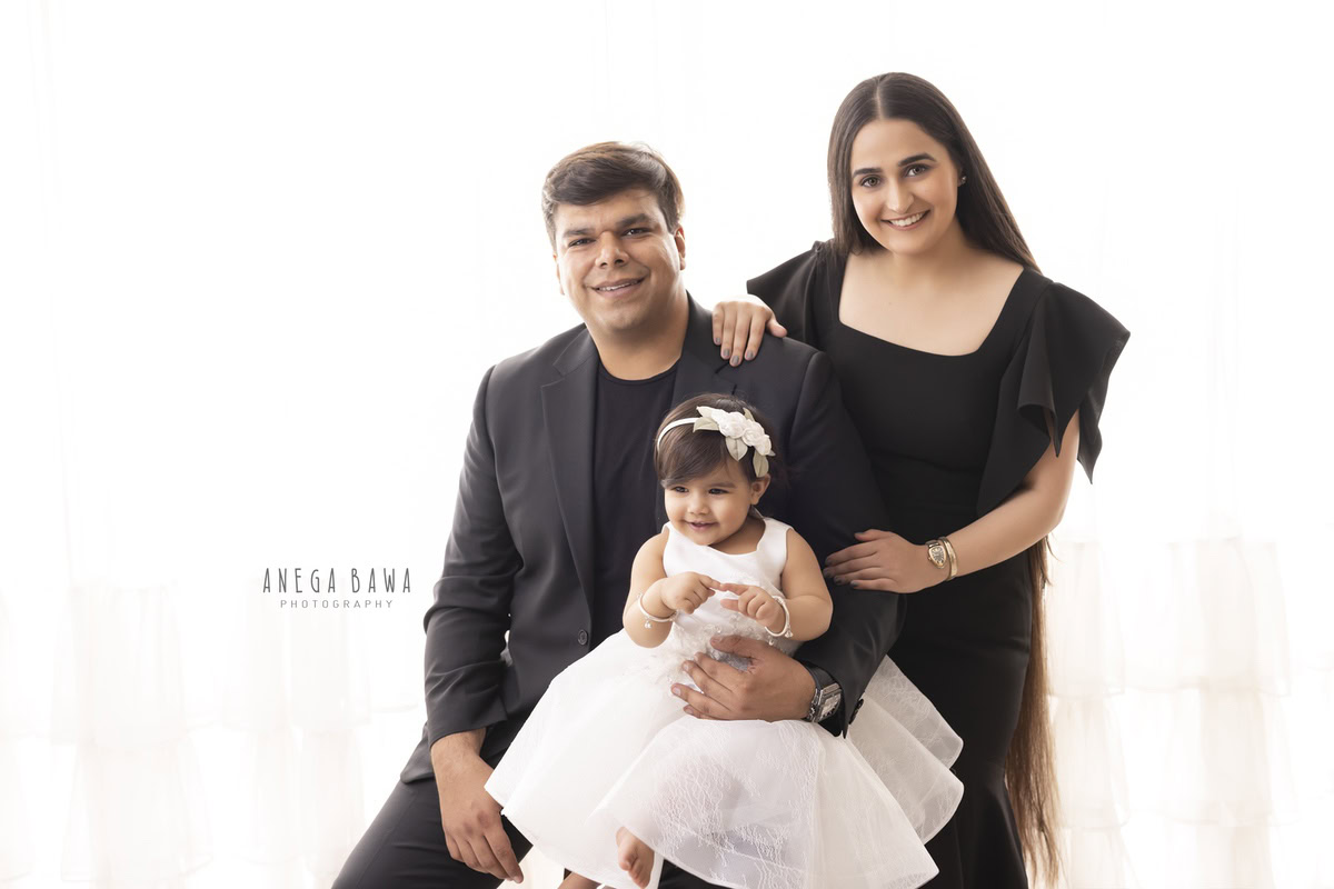 Charming 1-year-old girl seated on dad's lap, joyfully posing with mom against a pristine white backdrop. Captured by Anega Bawa Photography, specializing in family photoshoots in Gurgaon (Delhi NCR).