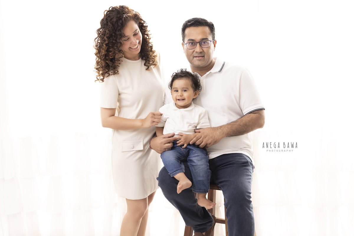 Adorable 1-year-old girl seated on dad's lap, posing with mom against a clean white backdrop. Photographed by Anega Bawa Photography, known for exquisite family photoshoots in Gurgaon (Delhi NCR).