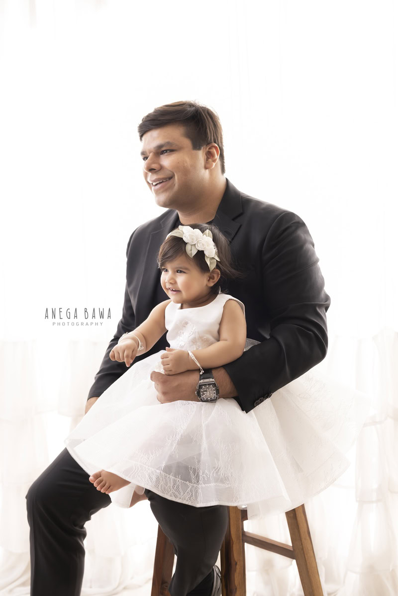 1-year-old girl seated on dad's lap against a serene white backdrop. Captured by Anega Bawa Photography in Gurgaon (Delhi NCR).
