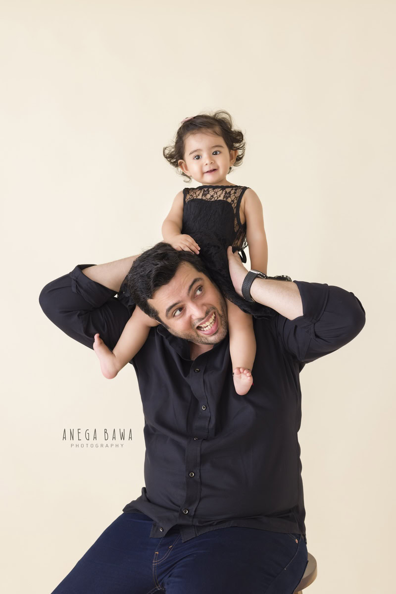 1-year-old girl seated on dad's shoulders against a serene beige backdrop. Captured by Anega Bawa Photography, Gurgaon (Delhi, NCR).