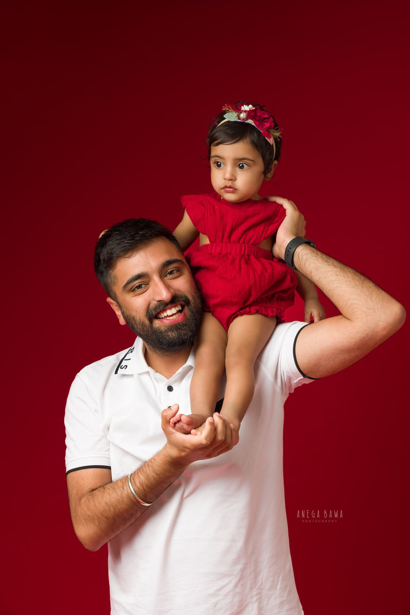 1-year-old girl seated on dad's shoulders, wearing a red dress, against a red backdrop, family photoshoot by Anega Bawa, Gurgaon (Delhi, NCR)