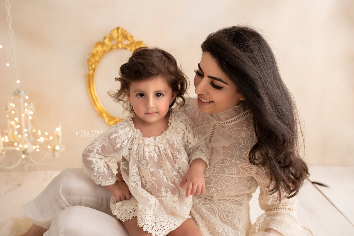 1-year-old girl seated on the floor on mom's lap, against a beige backdrop with a golden frame, wearing a net top, family photoshoot by Anega Bawa, Gurgaon (Delhi, NCR)