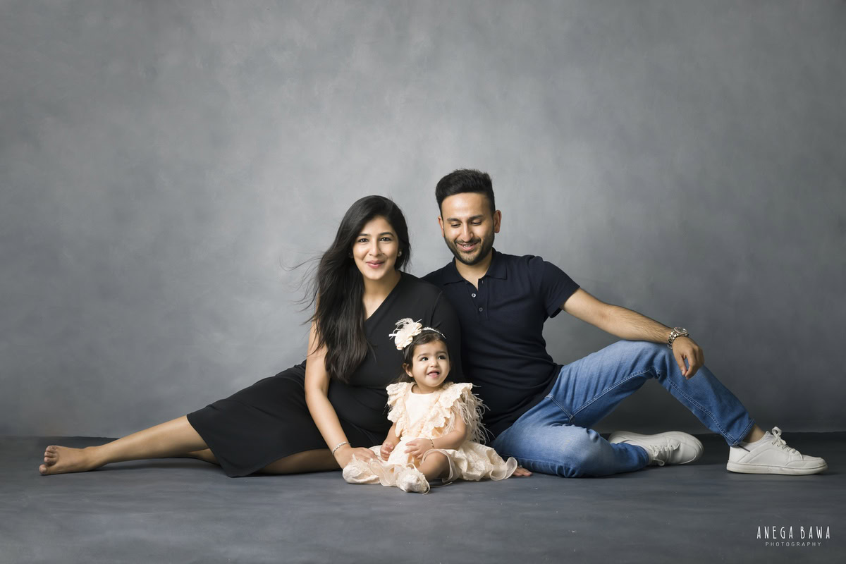 1-year-old girl seated on the floor, posing with mom and dad against a grey backdrop, family photoshoot by Anega Bawa, Gurgaon (Delhi, NCR)