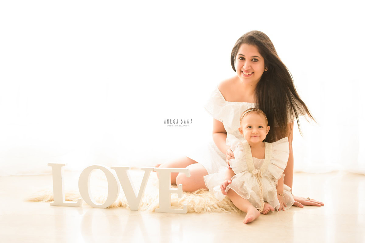 1-year-old girl seated on the floor with mom, on a beige rug against a white backdrop, family photoshoot by Anega Bawa, Gurgaon (Delhi, NCR)