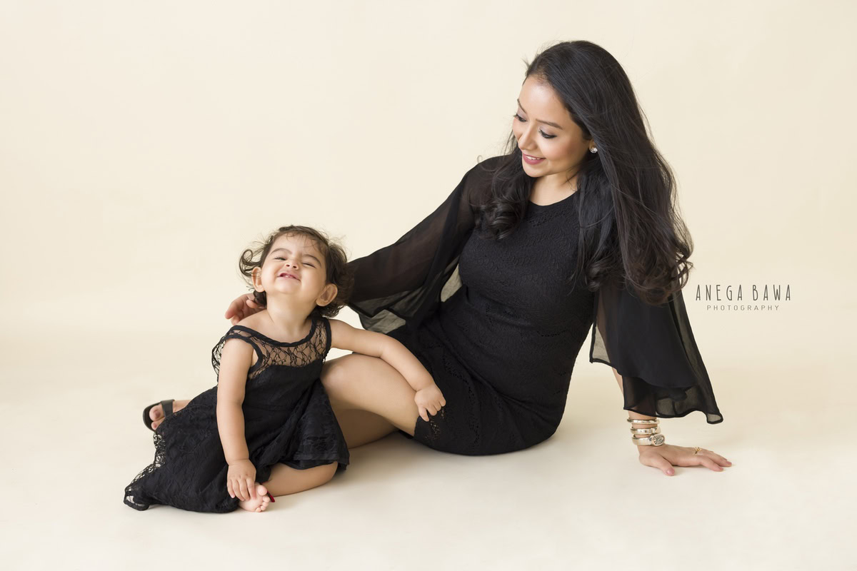 1-year-old girl seated on the floor with mom, striking a cute pout pose against a beige backdrop, captured by Anega Bawa, family photographer in Gurgaon (Delhi NCR).