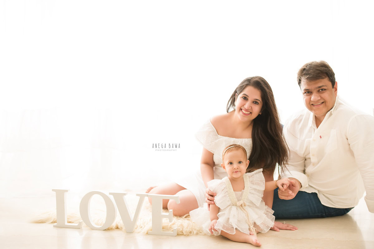 1-year-old girl seated on the floor with mom and dad, on a beige rug against a white backdrop. Captured by Anega Bawa Photography, Gurgaon (Delhi, NCR).