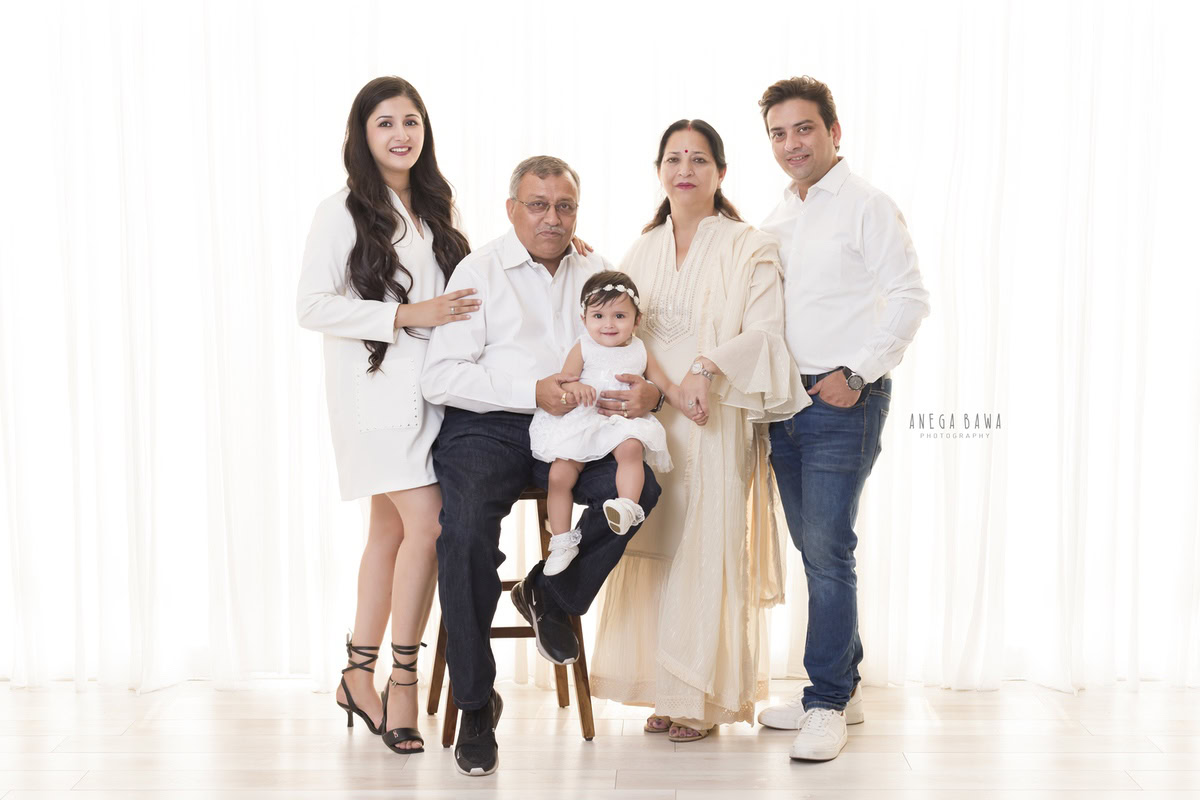 1-year girl seated on grandfather's lap, surrounded by grandma, mom, and dad in a family pose against a white backdrop. Captured by Anega Bawa Photography in Gurgaon (Delhi NCR).