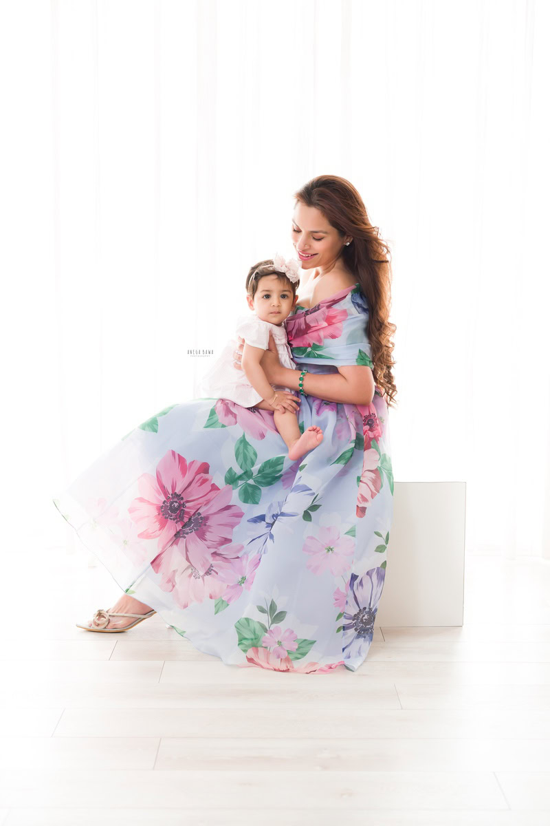 1-year-old girl seated on mom's lap in an adorable pose against a white backdrop. Captured by Anega Bawa Photography, Gurgaon (Delhi, NCR).