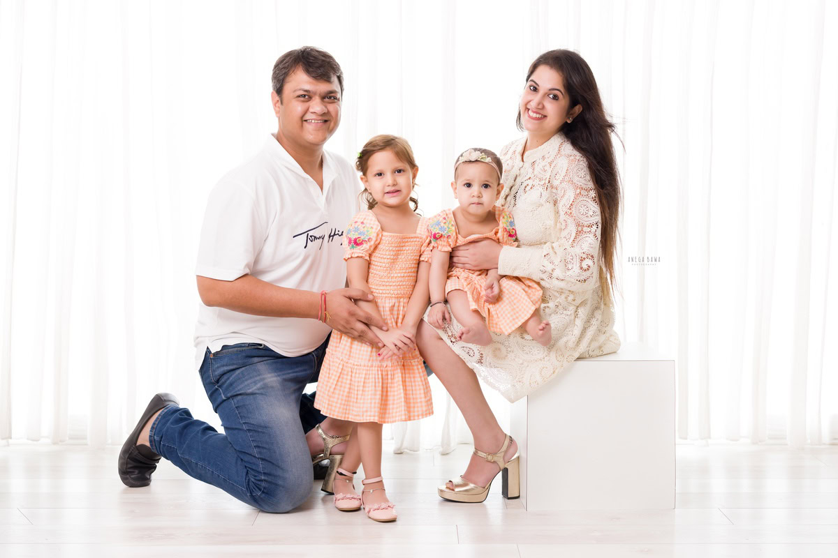 1-year-old girl seated on her mom's lap, accompanied by her dad and elder sister, posing for a family portrait against a serene white backdrop. Capture this heartwarming moment with Anega Bawa Photography, specializing in family photoshoots in Gurgaon (Delhi NCR).
