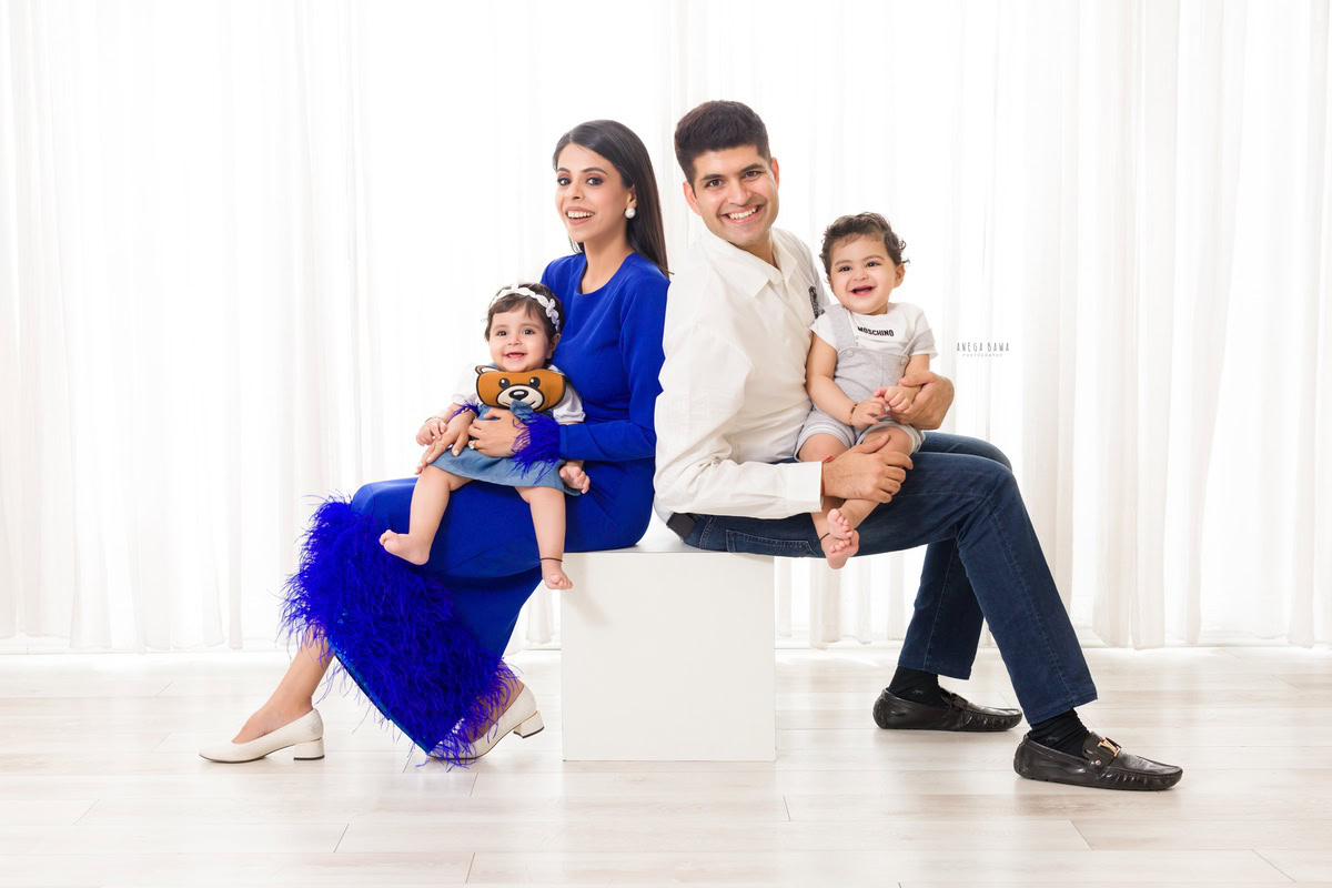 1-year girl seating on mom's lap, with elder brother and dad in a family pose against a white backdrop, captured by Anega Bawa Photography for a photoshoot in Gurgaon (Delhi NCR).