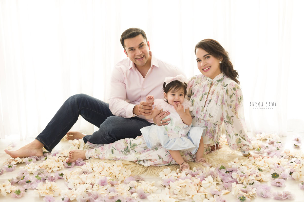 1-year girl seating on mom's lap on floor with dad, beige rug and flowers on floor, white backdrop, captured by Anega Bawa Photography in Gurgaon (Delhi NCR).