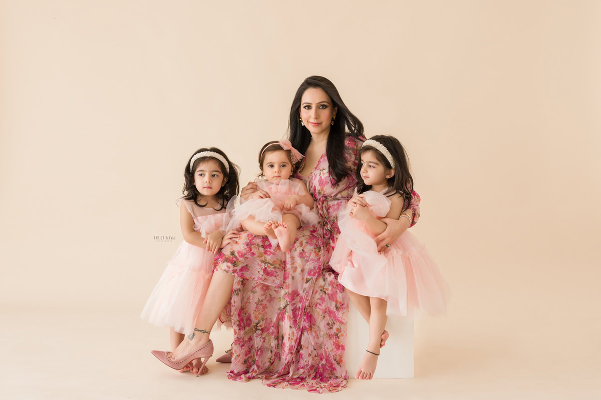 1-year-old girl seated on mom's lap, posing with two elder sisters against a beige backdrop, family photoshoot by Anega Bawa, Gurgaon (Delhi, NCR)