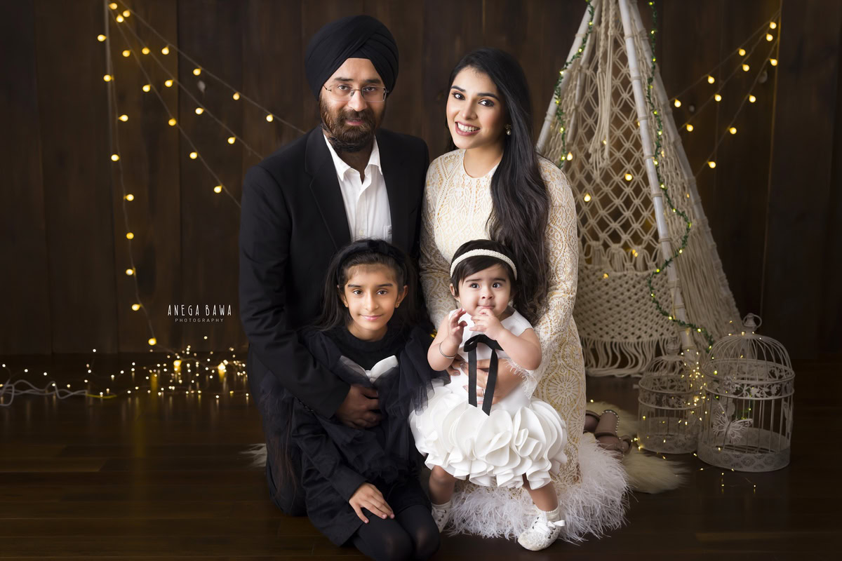 1-year-old girl seated on mom's lap, posing with elder sister and dad against a brown backdrop with fairy lights and castles, family photoshoot by Anega Bawa, Gurgaon (Delhi, NCR)