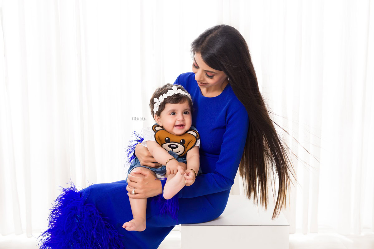 1-year-old girl seated on mom's lap, wearing a cute headband, against a white backdrop, family photoshoot by Anega Bawa, Gurgaon (Delhi, NCR)