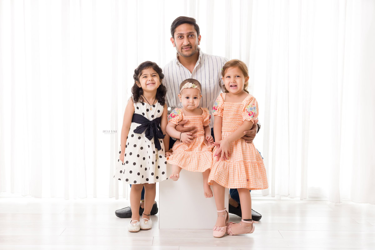 1-year girl seating on stool, posing with dad and two elder sisters against a white backdrop, captured by Anega Bawa Photography in Gurgaon (Delhi NCR).