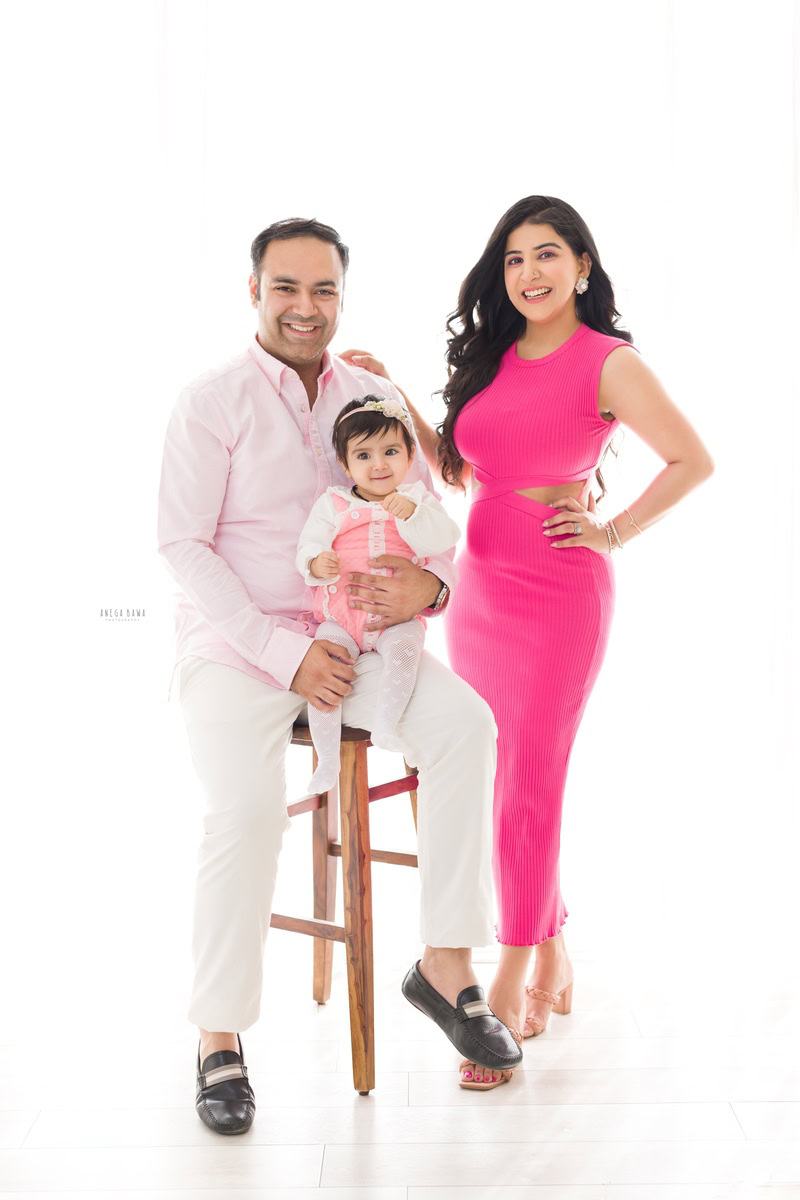 1-year girl seated on a stool with dad, while mom stands beside her against a serene white backdrop, captured by Anega Bawa Photography in Gurgaon (Delhi NCR).