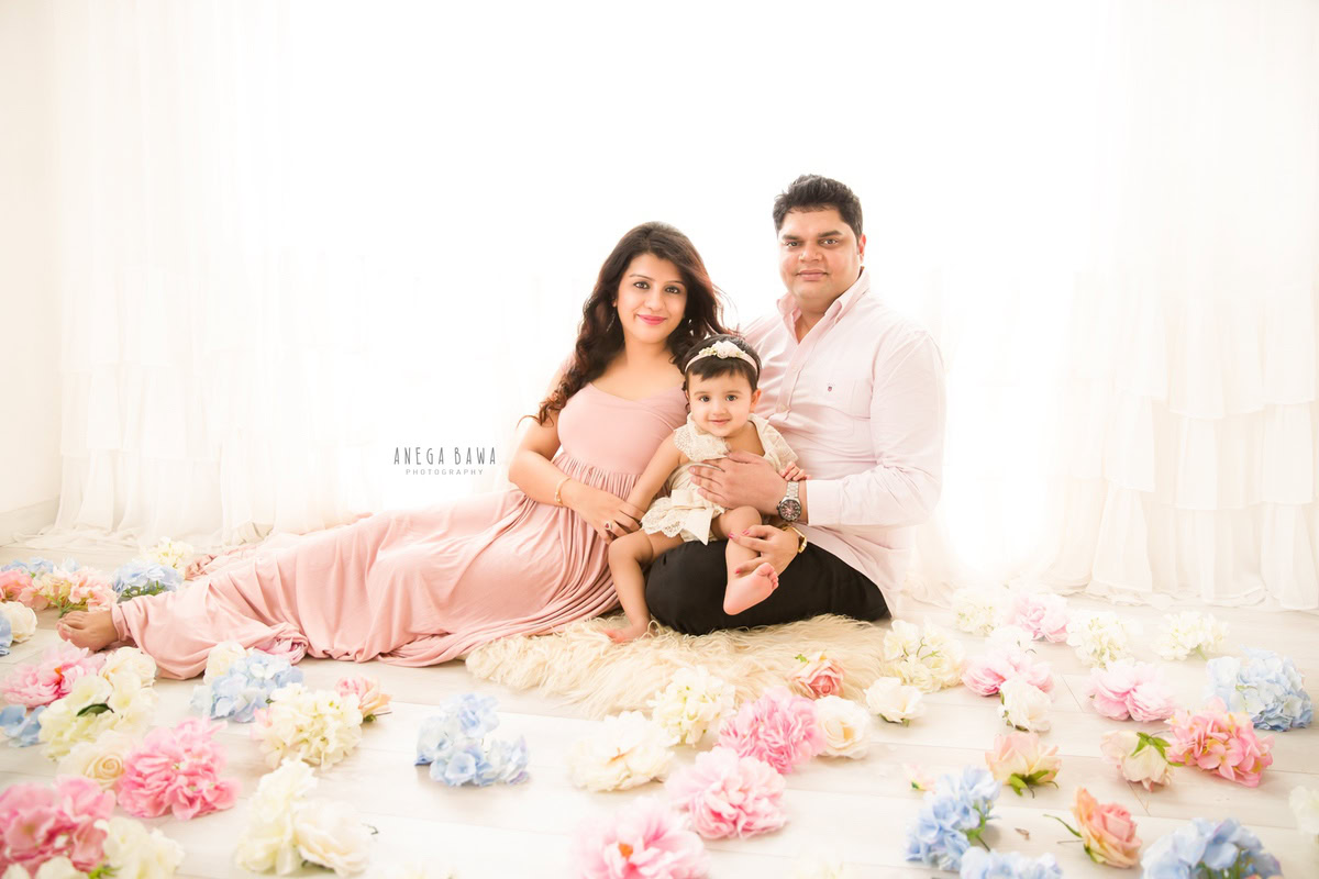 1-year-old girl seated with mom and dad on the floor surrounded by flowers against a white backdrop. Captured by Anega Bawa Photography, Gurgaon (Delhi, NCR).