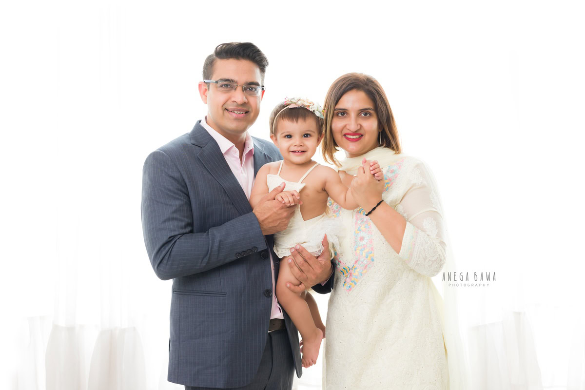 1-year-old girl smiling with mom and dad in a cute pose against a white backdrop, captured by Anega Bawa, family photographer in Gurgaon (Delhi NCR).