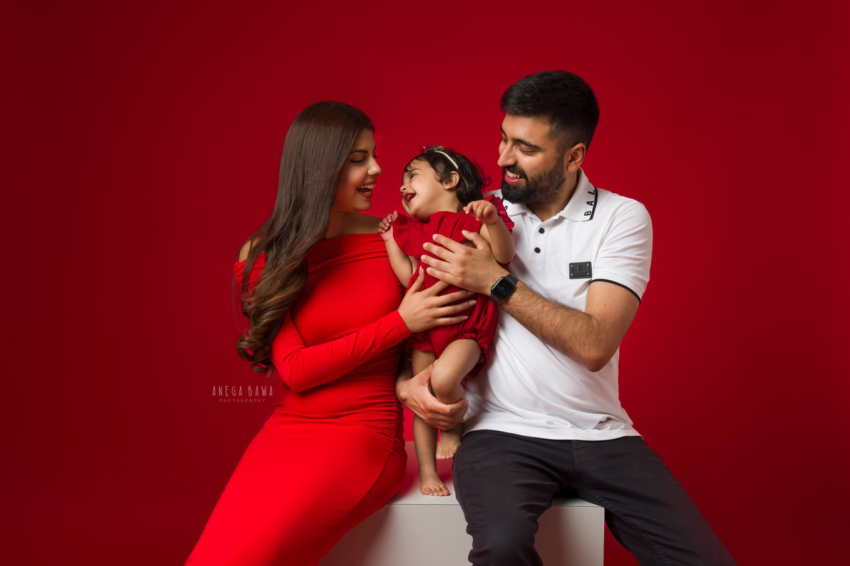 1-year girl smiling warmly with mom and dad against a vibrant red backdrop, photographed by Anega Bawa Photography in Gurgaon (Delhi NCR).