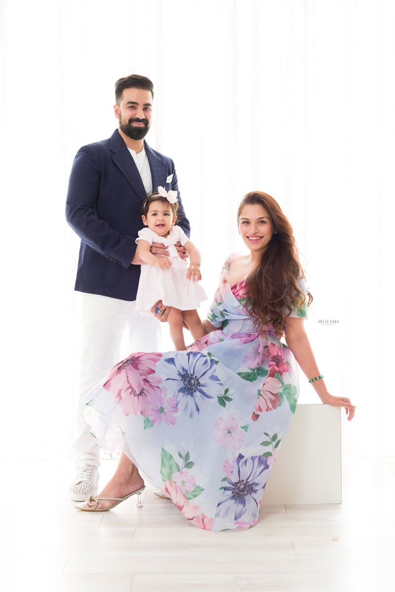 1-year-old girl standing on mom's lap with dad posing against a white backdrop, captured by Anega Bawa, family photographer in Gurgaon (Delhi NCR).