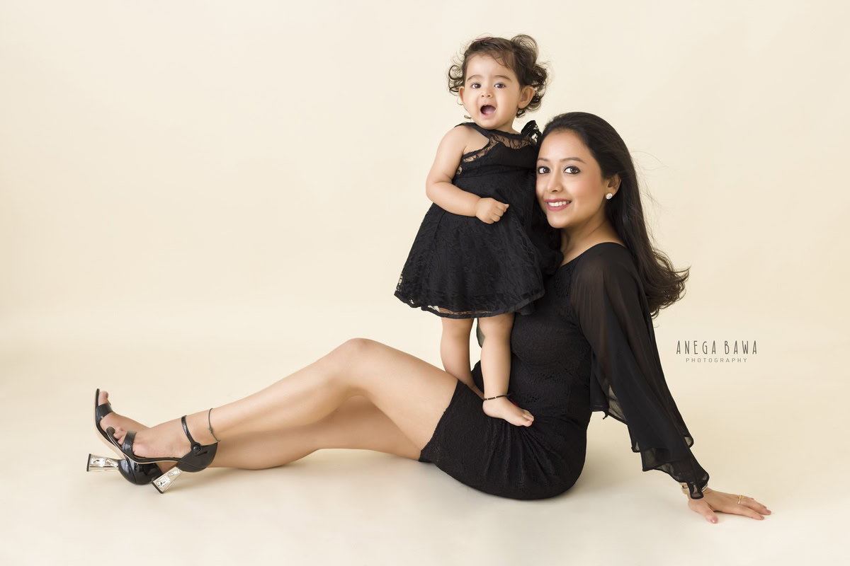 1-year-old girl standing on mom's lap, both twinning in black, against a beige backdrop. Captured by Anega Bawa Photography, Gurgaon (Delhi, NCR).
