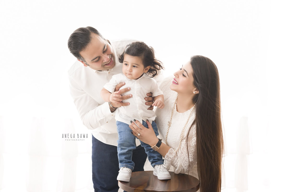 1-year-old girl standing on a stool, posing with mom and dad against a neutral backdrop, beautifully captured by Anega Bawa Photography in Gurgaon (Delhi NCR).