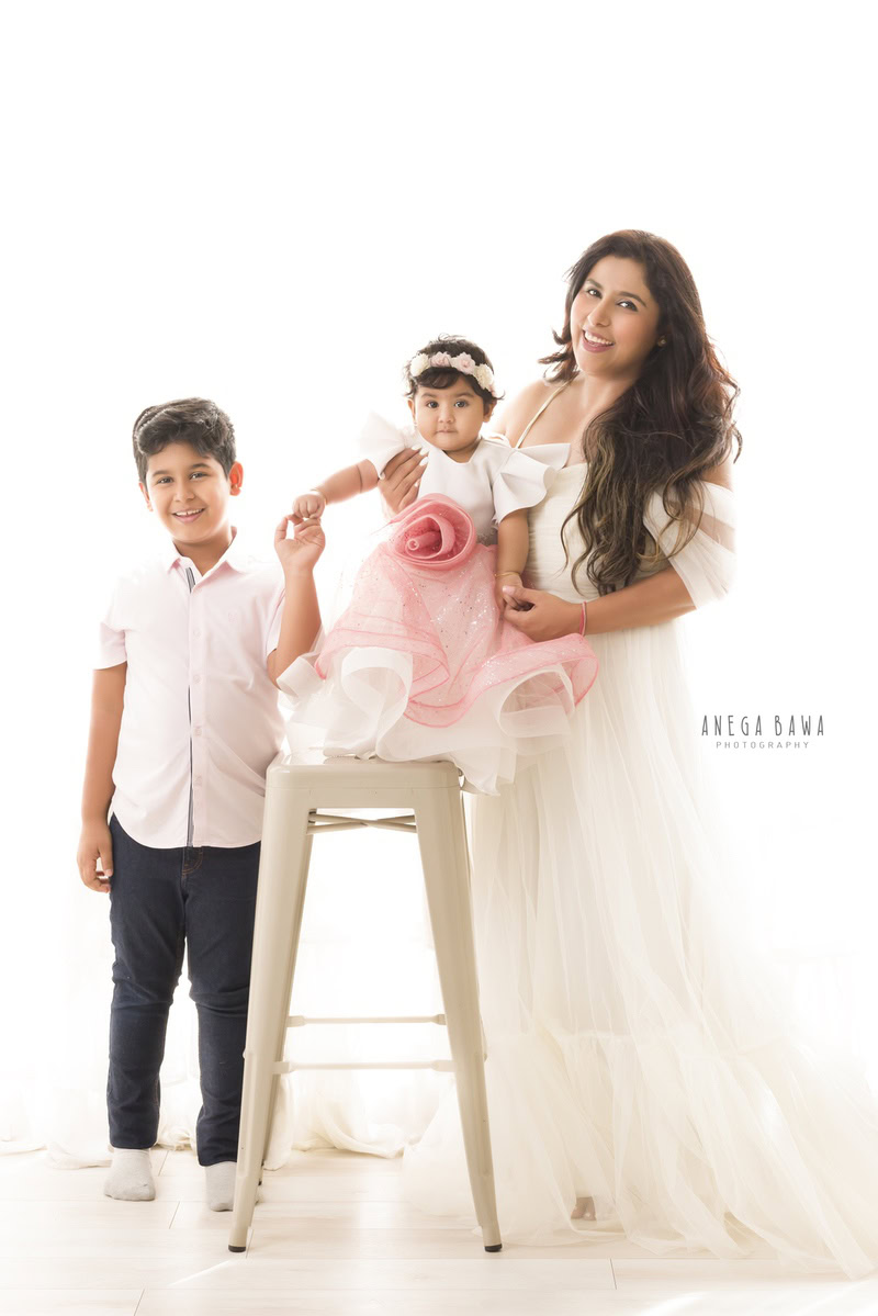 1-year-old girl in a pink dress standing on a stool, posing with mom and elder brother against a serene white backdrop, captured by Anega Bawa Photography in Gurgaon (Delhi NCR).