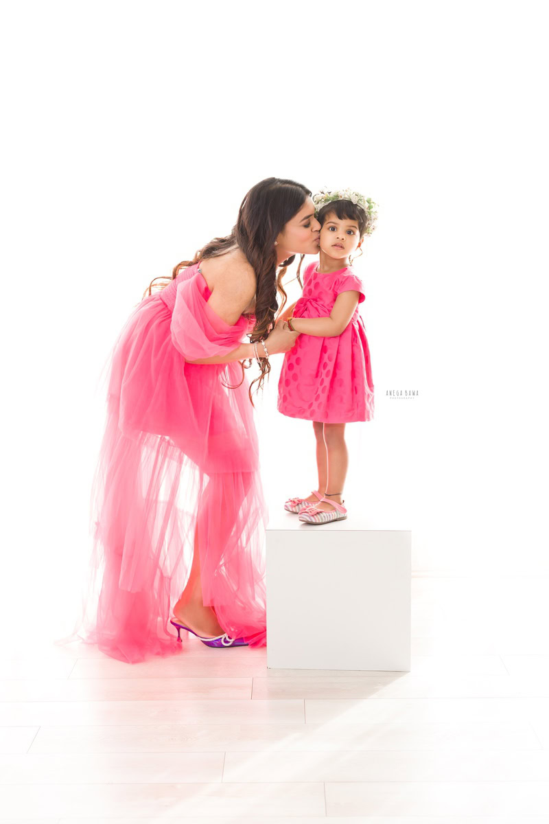 1-year-old girl standing on a white stool with mom in a kissing pose against a white backdrop, captured by Anega Bawa, family photographer in Gurgaon (Delhi NCR).