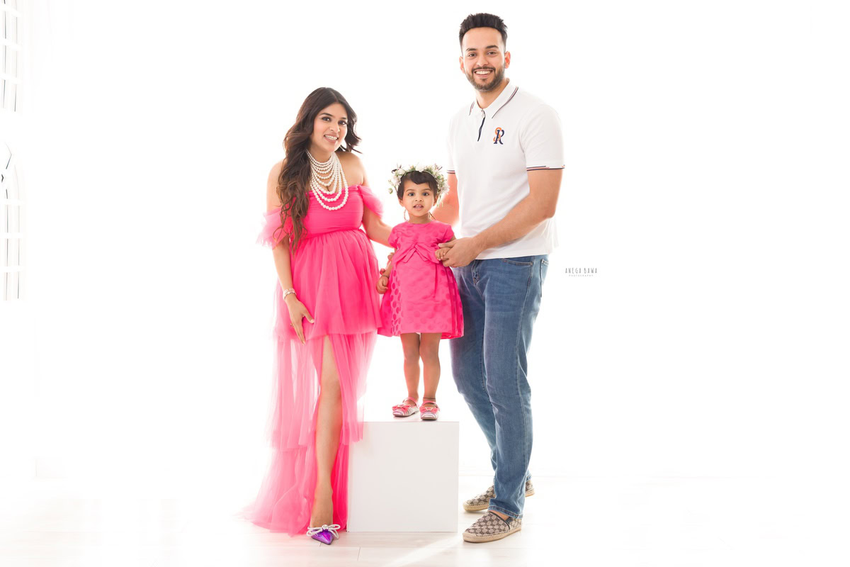 1-year-old girl standing on a white stool, twinning with mom who is wearing a hot pink dress, posing with dad against a white backdrop, captured by Anega Bawa, family photographer in Gurgaon (Delhi NCR).
