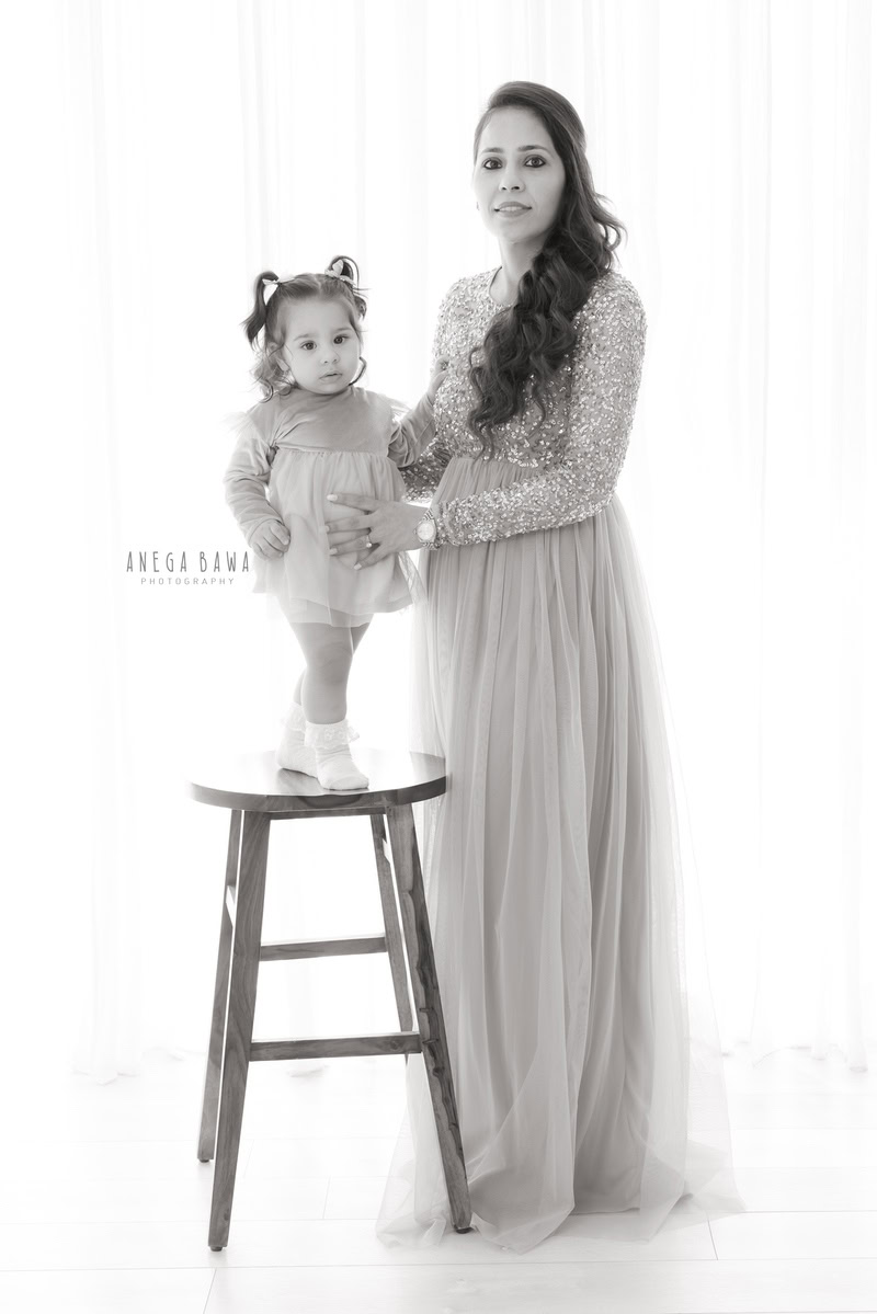 1-year girl standing on a wooden stool in a cute beige dress, posing with mom against a serene white backdrop, captured by Anega Bawa Photography in Gurgaon (Delhi NCR).