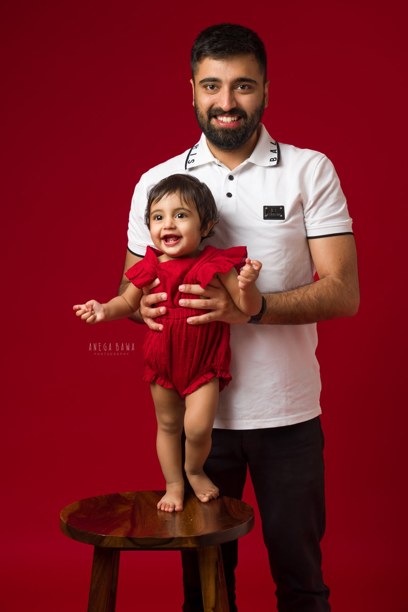 1-year-old girl standing on a wooden stool, held by dad, against a vibrant red backdrop. Captured by Anega Bawa Photography, Gurgaon (Delhi, NCR).
