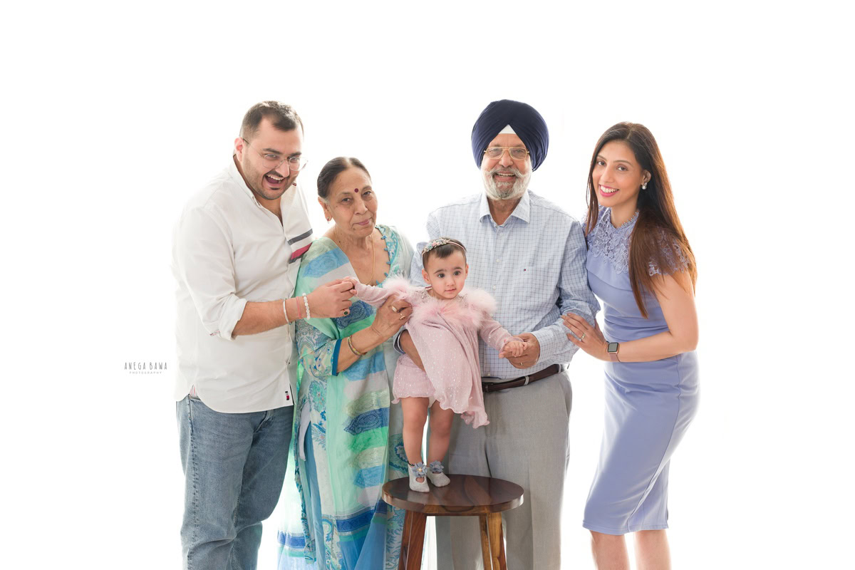 1-year-old girl standing on a wooden stool with her mom, dad, and grandparents against a serene white backdrop, captured in a family pose by Anega Bawa Photography in Gurgaon (Delhi NCR).