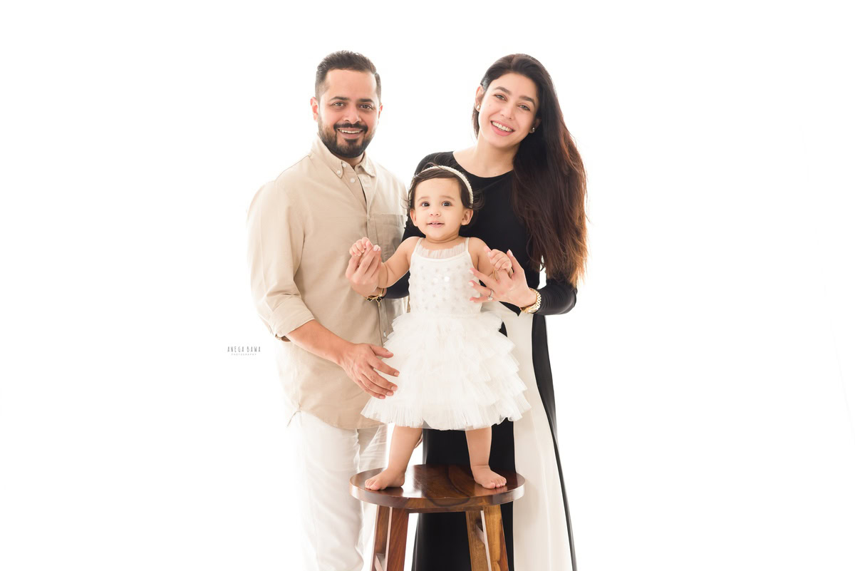 1-year-old girl standing on a wooden stool with mom and dad in the background against a serene white backdrop. Captured by Anega Bawa Photography, specializing in family photo shoots in Gurgaon (Delhi NCR).