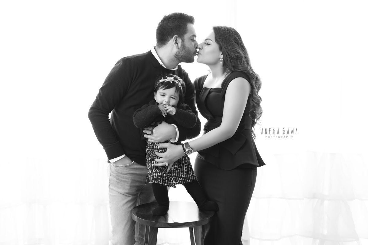 1-year-old girl standing on a wooden stool with mom and dad in a kissing pose against a white backdrop, capturing a cute family pose, by Anega Bawa, family photographer in Gurgaon (Delhi NCR).