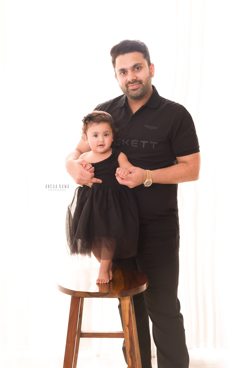 1-year-old girl standing on a wooden stool, posing with dad against a clean white backdrop. Captured by Anega Bawa Photography, Gurgaon (Delhi, NCR).