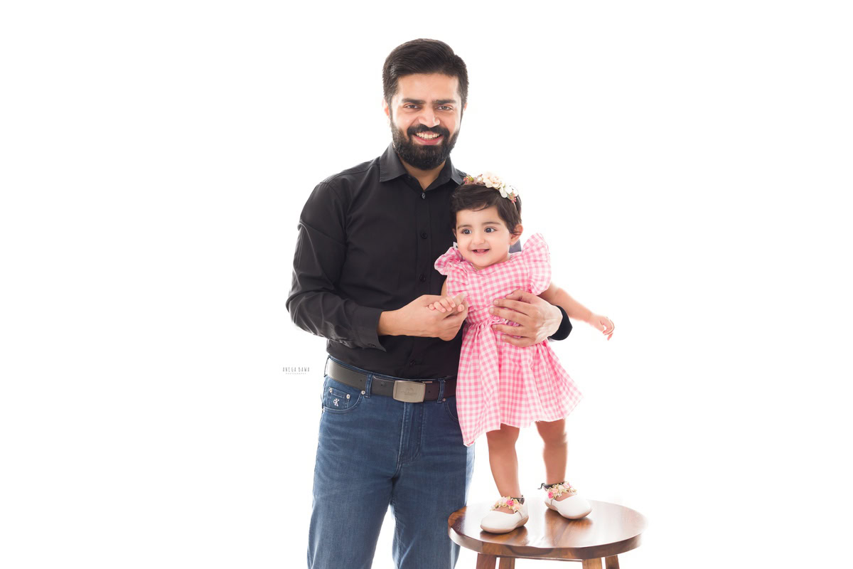 1-year-old girl standing on a wooden stool, posing with dad against a white backdrop, captured by Anega Bawa, family photographer in Gurgaon (Delhi NCR).
