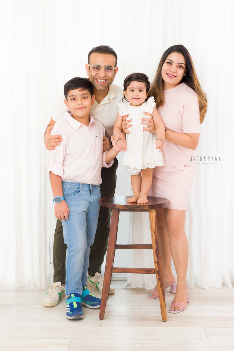 1-year-old girl standing on a wooden stool, posing with her elder brother, mom, and dad against a white backdrop, captured beautifully by Anega Bawa Photography for a family photoshoot in Gurgaon (Delhi NCR).