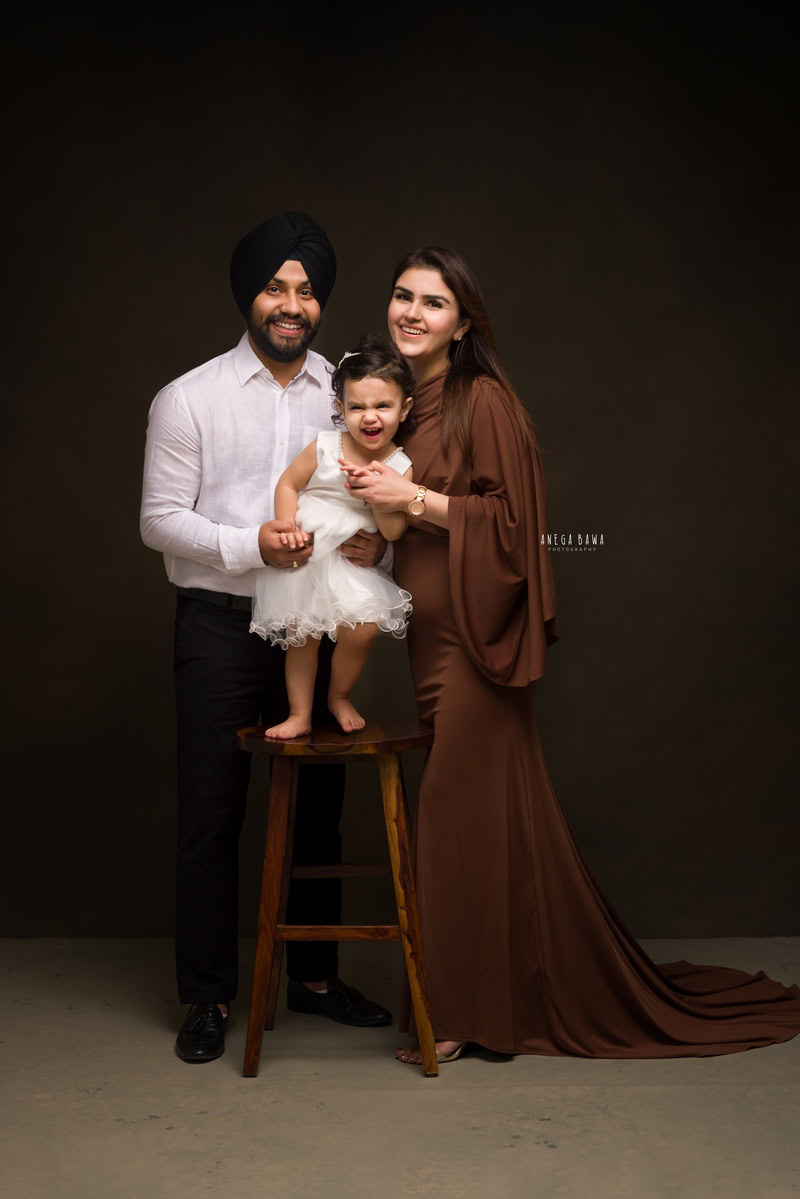 1-year-old girl standing on a wooden stool, posing with mom and dad against a brown backdrop, captured by Anega Bawa, family photographer in Gurgaon (Delhi NCR).