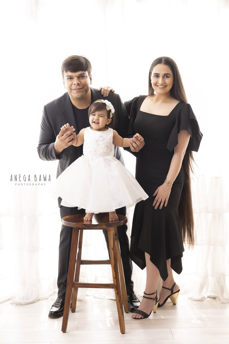 -year-old girl standing on a wooden stool, posing with mom and dad against a white backdrop, captured by Anega Bawa, family photographer in Gurgaon (Delhi NCR)