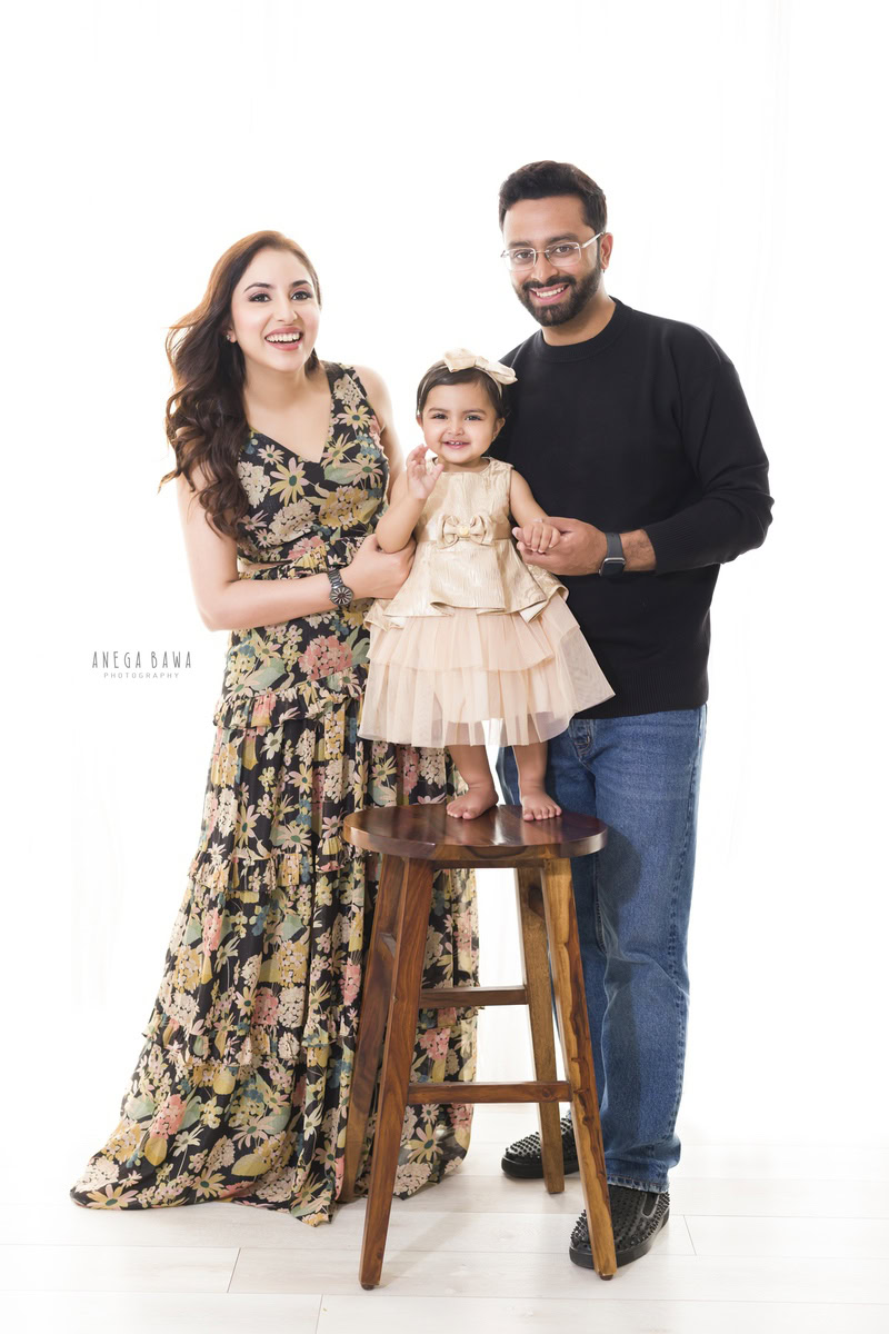 1-year-old girl standing on a wooden stool, posing with mom and dad against a white backdrop, family photoshoot by Anega Bawa, Gurgaon (Delhi, NCR)