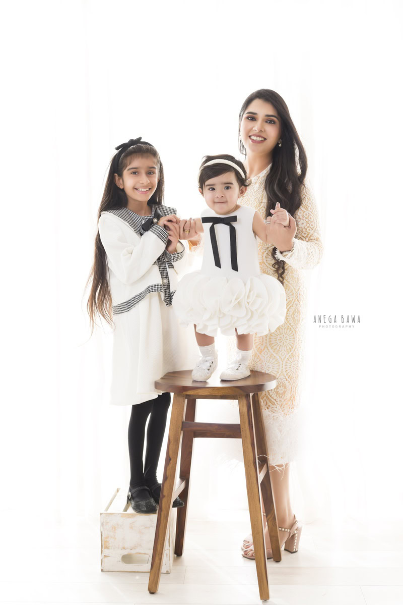 1-year-old girl standing on a wooden stool, posing with her mom and elder sister against a serene white backdrop, beautifully captured by Anega Bawa Photography for a family photoshoot in Gurgaon (Delhi NCR).