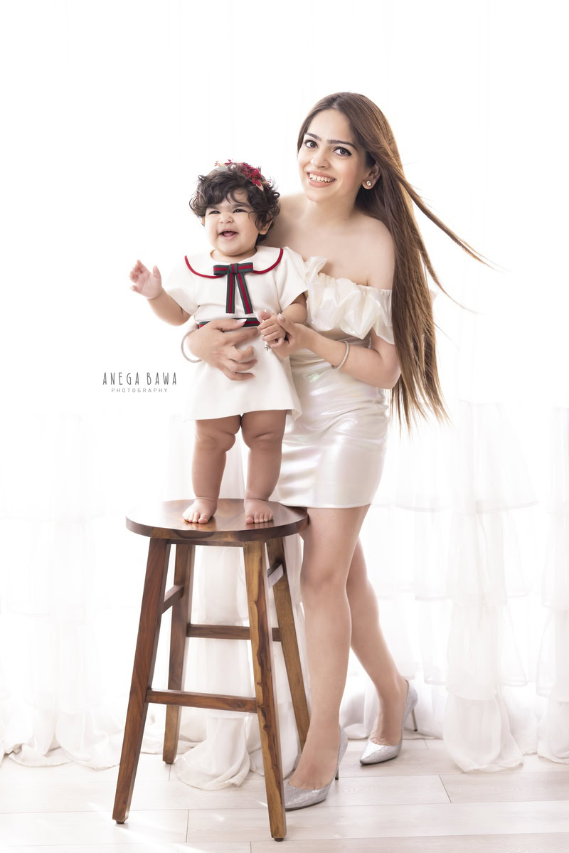1-year-old girl standing on a wooden stool, posing with mom against a serene white backdrop, beautifully captured by Anega Bawa Photography in Gurgaon (Delhi NCR).