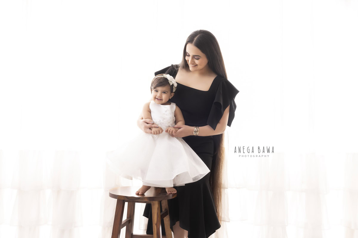1-year-girl standing on a wooden stool, posing with her mom against a serene white backdrop, captured by Anega Bawa Photography in Gurgaon (Delhi NCR).