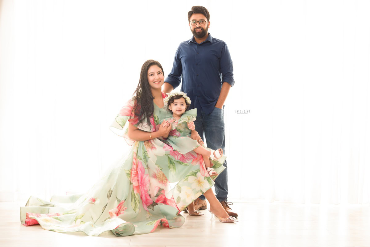 1-year-old girl elegantly twinning with mom in a vibrant floral dress and sparkling tiara band, joyfully posing with dad against a pristine white backdrop. Captured by Anega Bawa Photography, Gurgaon (Delhi, NCR).