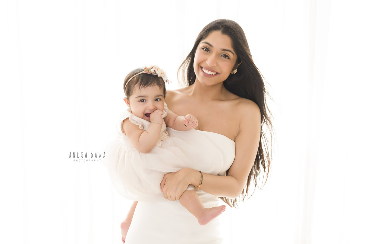 1-year-old girl wearing a cute headband, posing in mom's arms against a white backdrop, family photoshoot by Anega Bawa, Gurgaon (Delhi, NCR)