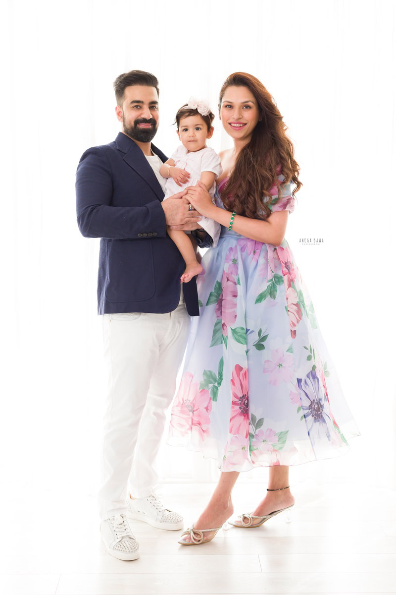 1-year-old girl wearing a cute headband, posing with mom and dad against a white backdrop, captured by Anega Bawa, family photographer in Gurgaon (Delhi NCR).