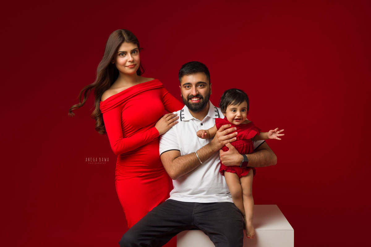 1-year-old girl wearing a cute red baby suit poses with mom and dad against a vibrant red backdrop, captured beautifully by Anega Bawa Photography for a family photoshoot in Gurgaon, Delhi NCR.