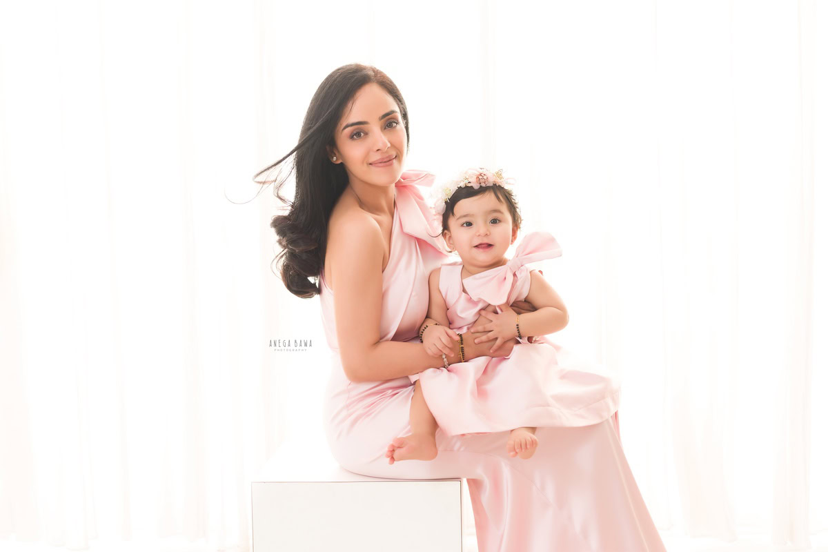 1-year-old girl wearing a pastel pink dress, seated on mom's lap against a white backdrop, family photoshoot by Anega Bawa, Gurgaon (Delhi, NCR)