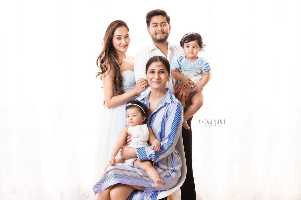 1-year-old twin girls seated on Granny's lap, with mom and dad in a family pose against a white backdrop, captured by Anega Bawa, family photographer in Gurgaon (Delhi NCR).