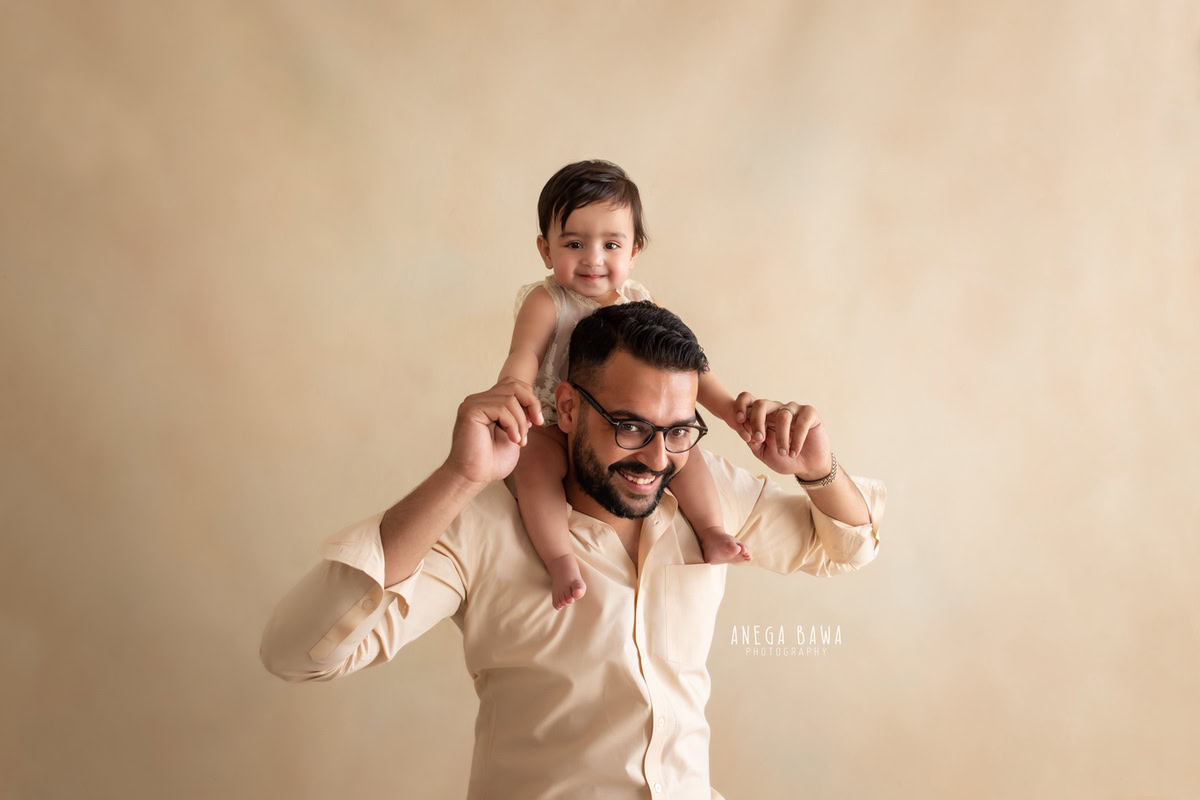 Playful 10-month-old boy posing on dad's shoulder against a soothing beige backdrop, captured beautifully by Anega Bawa in Gurgaon (Delhi, NCR)